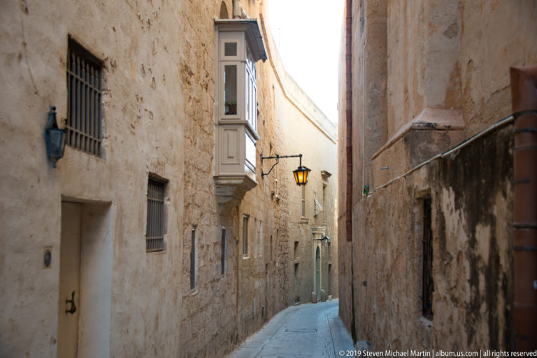 Narrow curving limestone lane with barred windows, wall lanterns, drainpipes, and a projecting enclosed balcony.