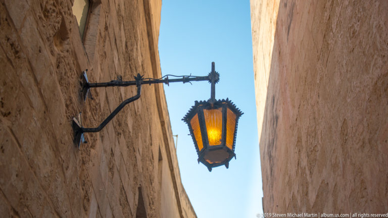 Amber lantern hangs from a decorative iron bracket between tall limestone walls beneath a narrow strip of blue sky.