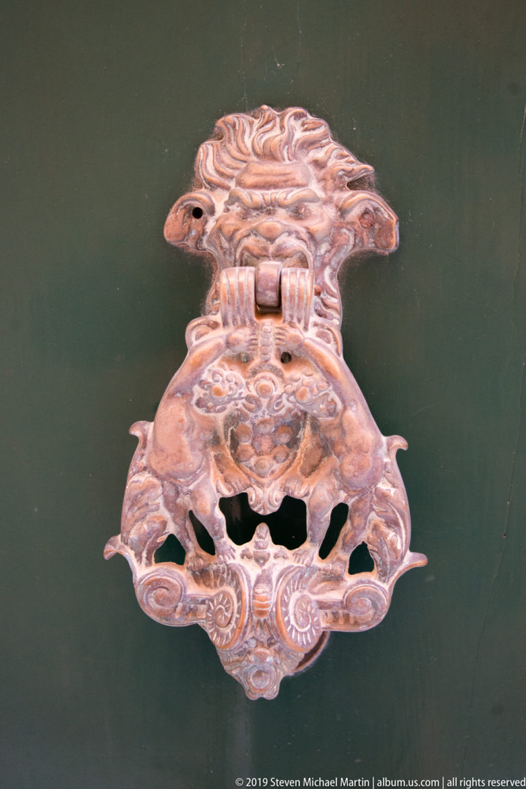 Ornate cast-metal door knocker with a sculpted face and pierced scrolling plate mounted on a dark door.