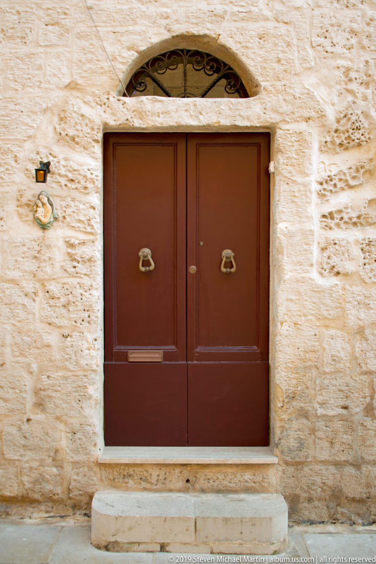 Reddish-brown double door with matching knockers and an arched fanlight set into a pale limestone wall.