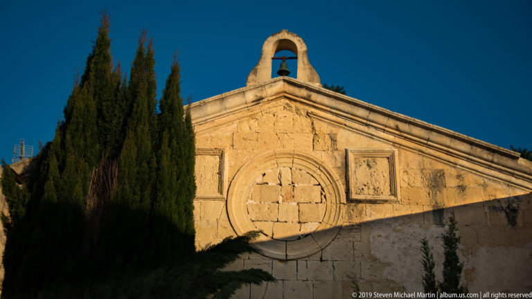 Sunlit limestone gable with a single bell, circular cross motif, and tall cypress trees against a blue sky.