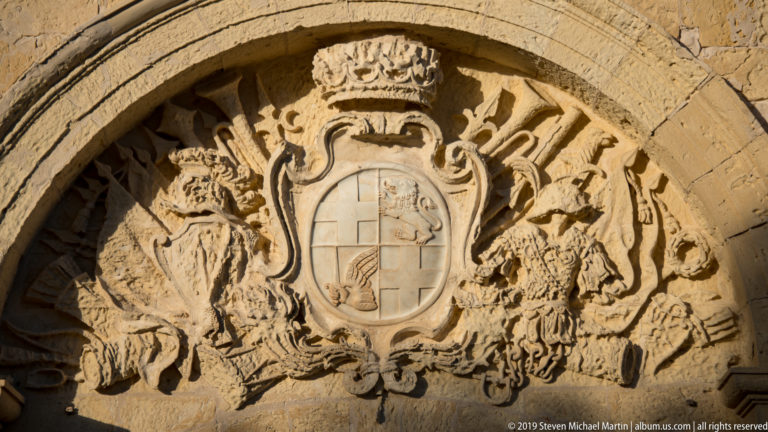 Weathered limestone coat of arms with crowned shield, two animal supporters, and elaborate scrollwork carved within an arch.