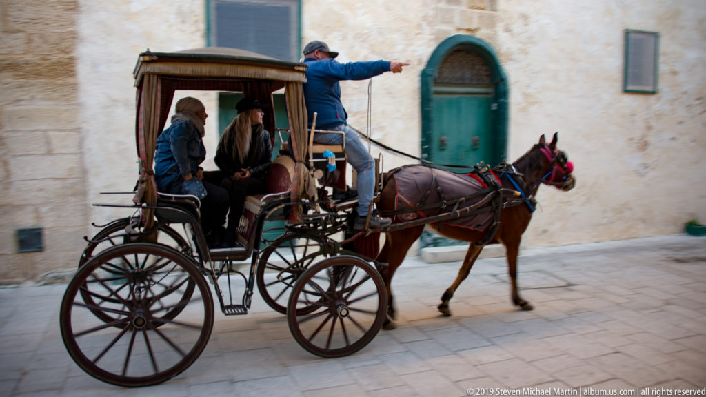 Horse-drawn carriage with passengers and driver moves past a green doorway on a pale limestone street in M’dina.