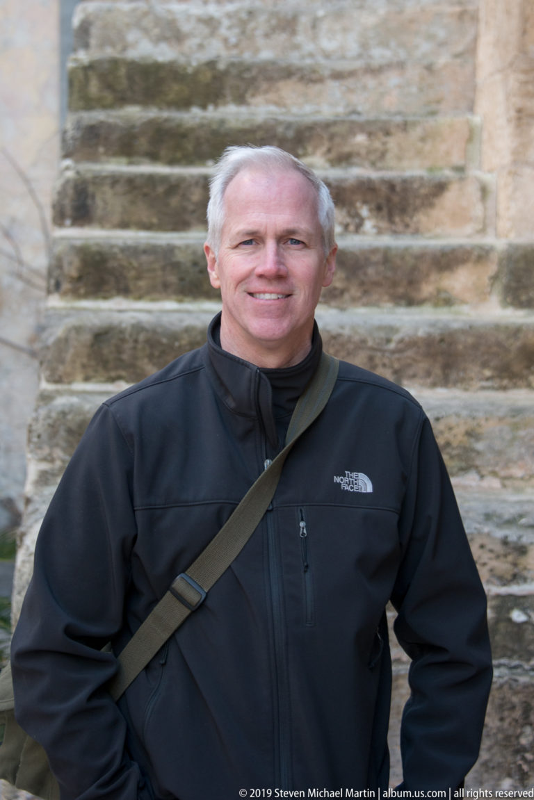 Smiling visitor in a dark jacket standing before weathered limestone steps.