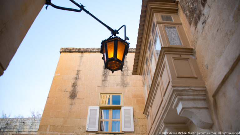 Hanging lantern beside a projecting enclosed balcony and shuttered window on limestone buildings.
