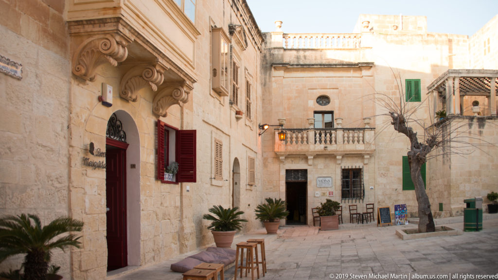 Small paved space between limestone façades with colorful shutters, balconies, potted plants, and café furniture.