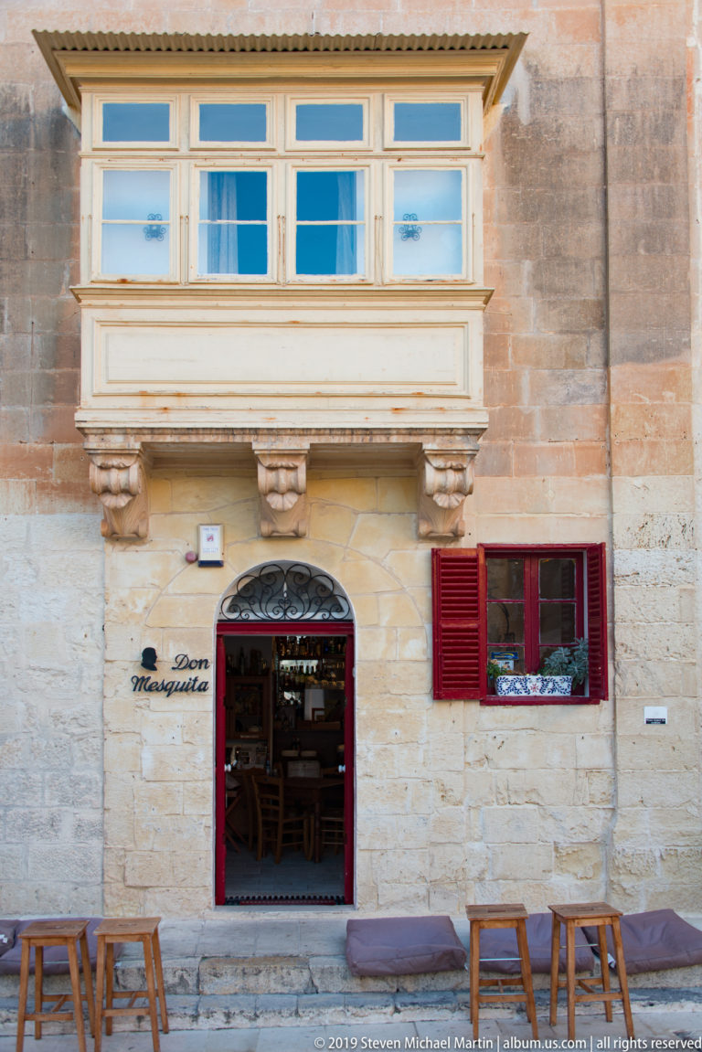 Café Marquit storefront with red trim, open doorway, pavement stools, and an enclosed wooden balcony on a limestone façade.