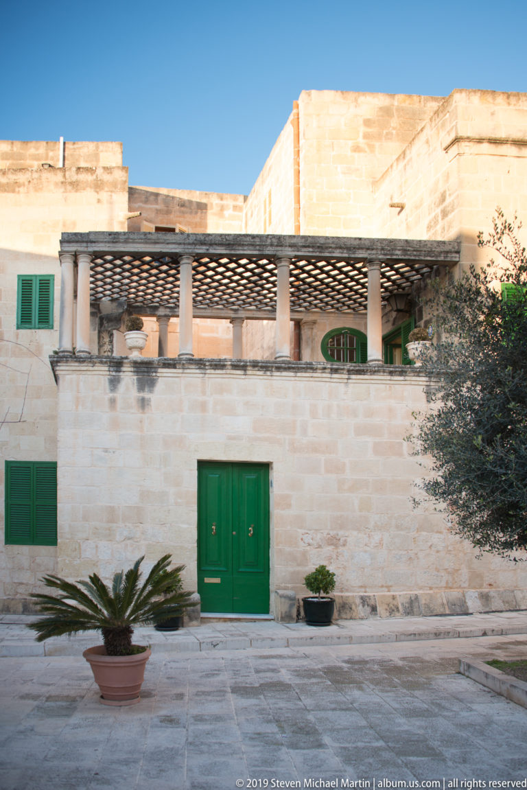 Low limestone courtyard façade with green doors and shutters, columned pergola, potted cycads, and olive tree.