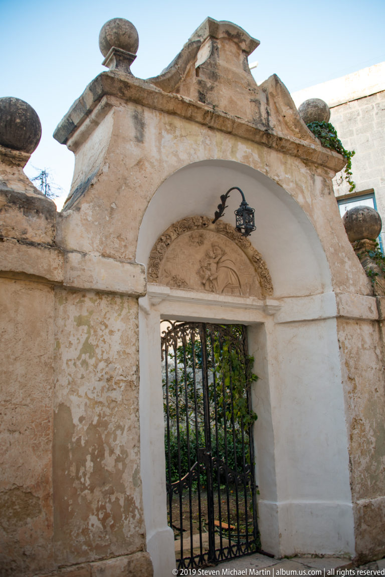 Weathered arched limestone entrance with black iron gate, broken pediment, painted recess, and greenery beyond.