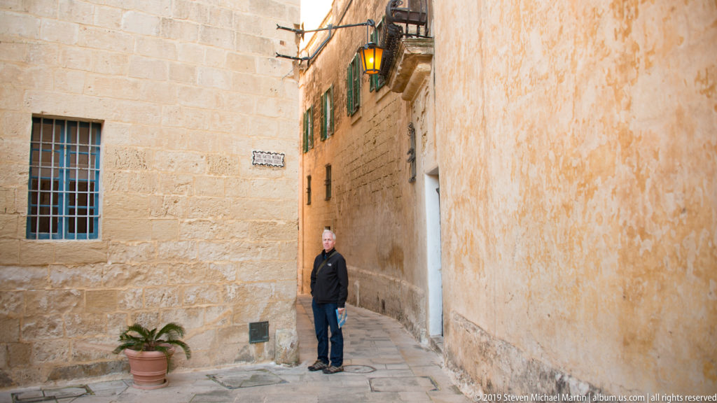 Visitor in dark clothing standing in a narrow limestone lane with barred window, potted plant, and lantern.