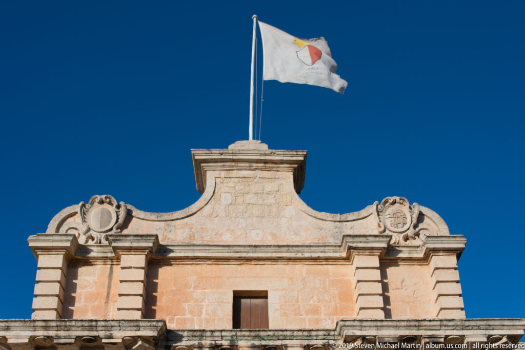 Maltese flag above a raised limestone parapet with carved heraldic roundels and rusticated corner details against blue sky.