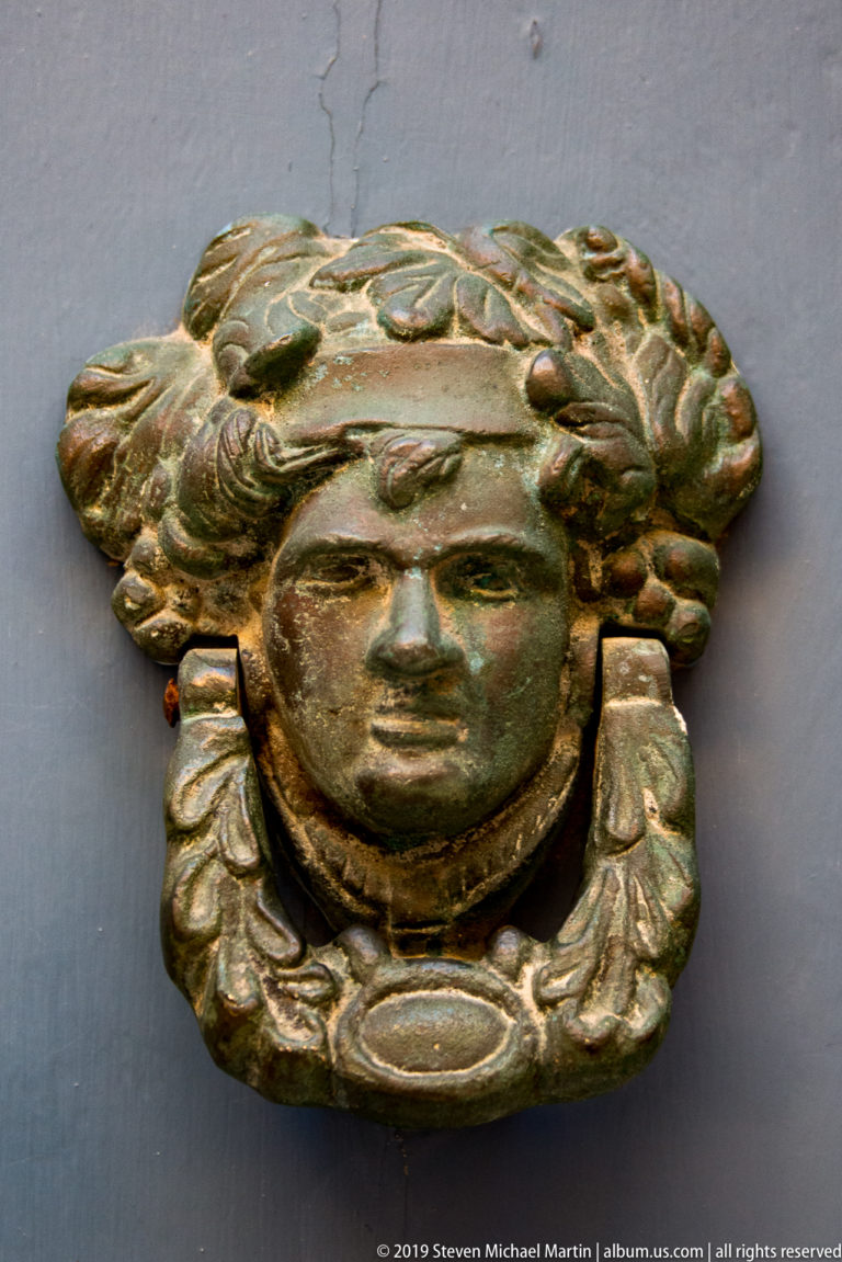 Metal door knocker shaped as a female head with flowing hair above a broad curved handle.