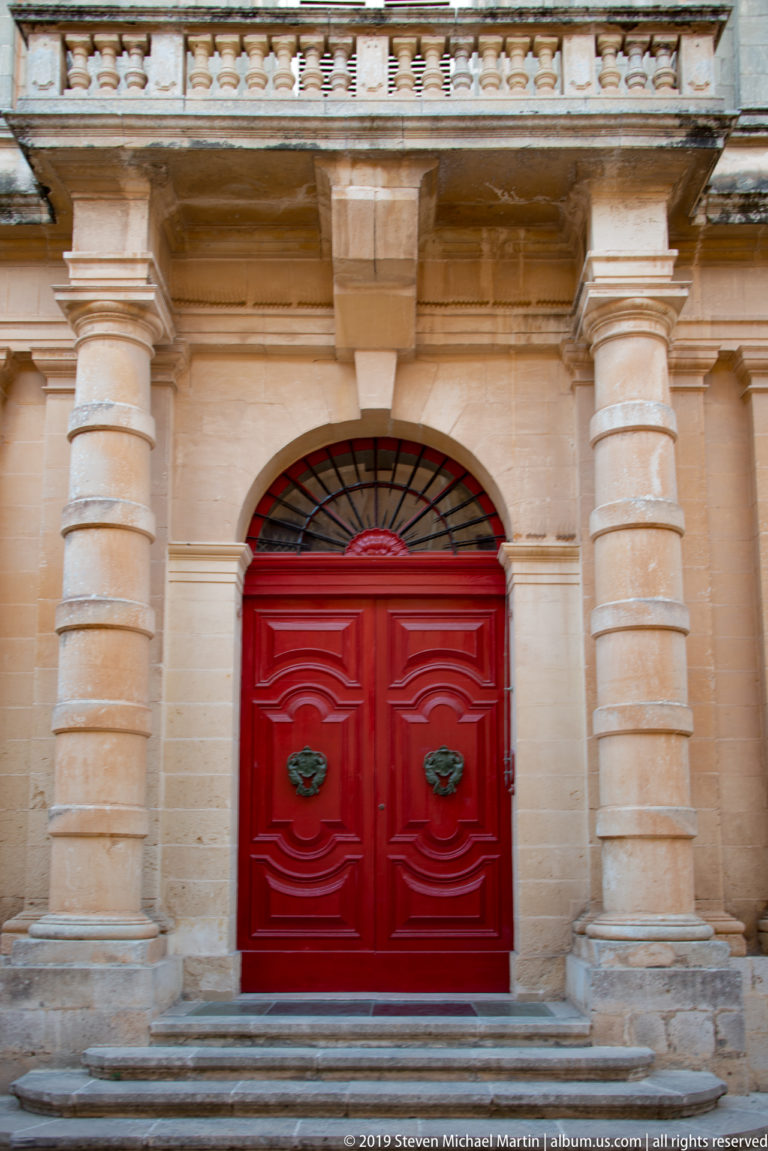Large red double door with sculptural knockers, semicircular fanlight, and rusticated limestone pilasters.