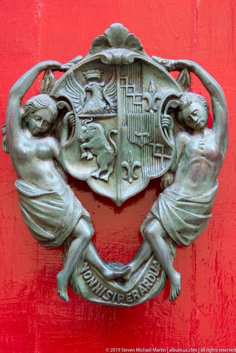 Heraldic metal door knocker with two supporting figures, central shield, and motto banner on a red door.