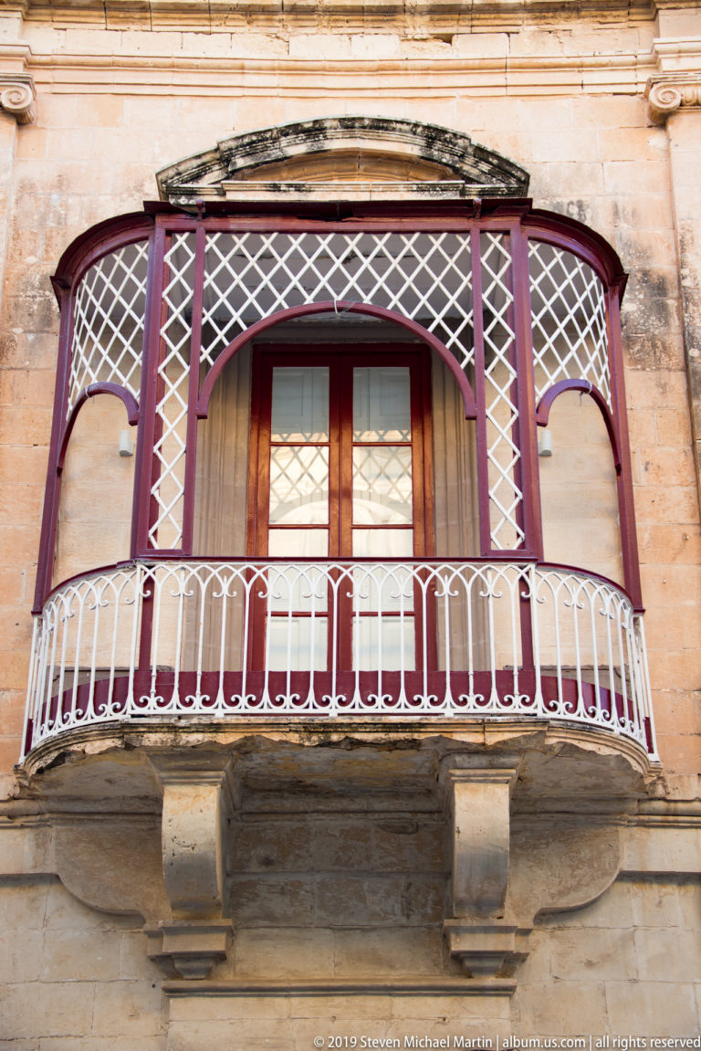 Deep red enclosed wooden balcony with lattice panels and white railing projecting from a limestone façade.