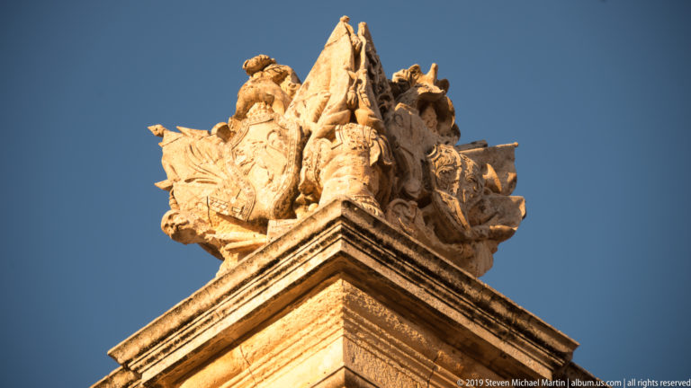 Elaborate carved stone finial with heraldic and trophy-like forms crowning a limestone corner against blue sky.