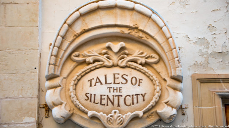 Decorative oval wall plaque reading “TALES OF THE SILENT CITY” within carved scroll and floral ornament.