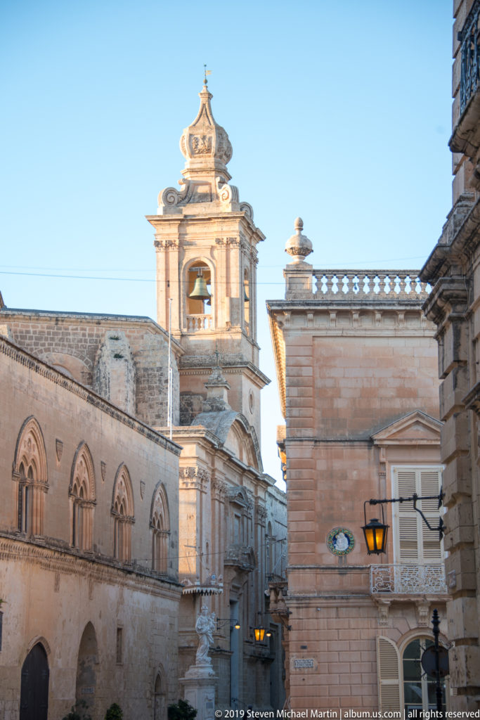 Ornate bell tower rises above pale limestone buildings with pointed arches, balustrades, shutters, and a street lantern.
