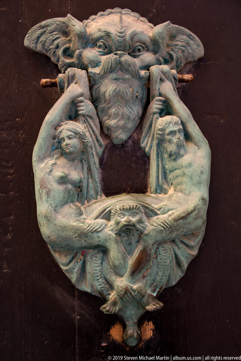 Ornate metal door knocker with a bearded face, scrolling moustache forms, and two long-necked birds.