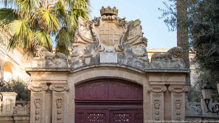 Red arched gate framed by carved limestone and topped by a large crowned coat of arms with scrolling supporters.