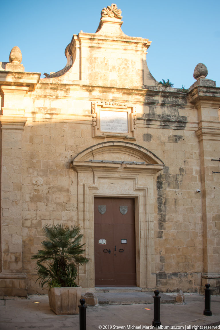 Weathered limestone façade with brown double doors, curved pediment, potted palm, and bollards.