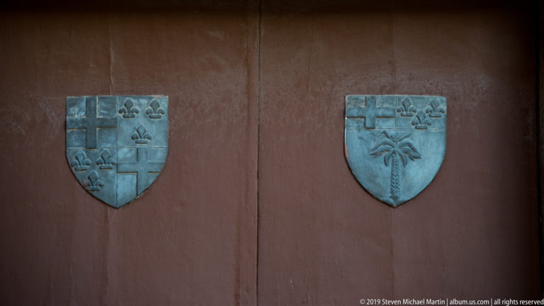 Two metal heraldic shields with crosses, fleurs-de-lis, and plant motifs mounted on a red door.
