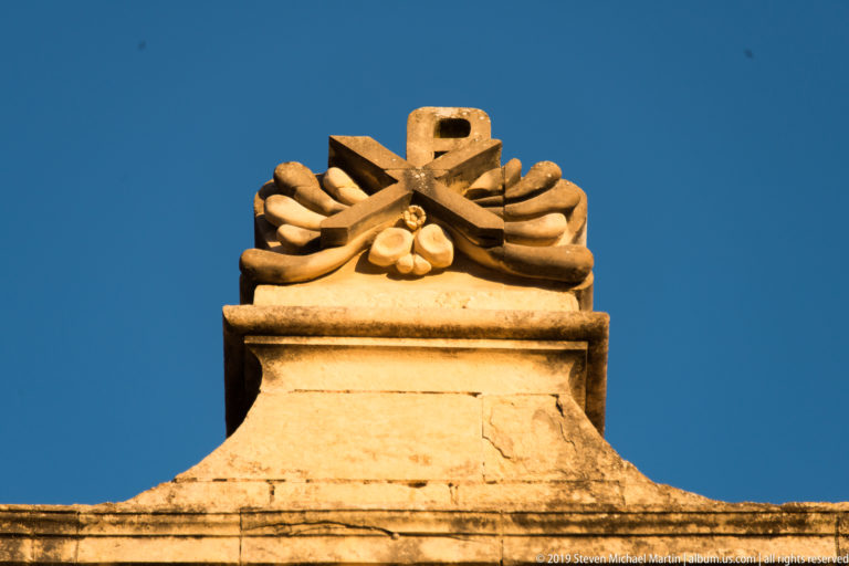 Symmetrical carved stone ornament resembling a tied bundle crowning a limestone parapet against blue sky.
