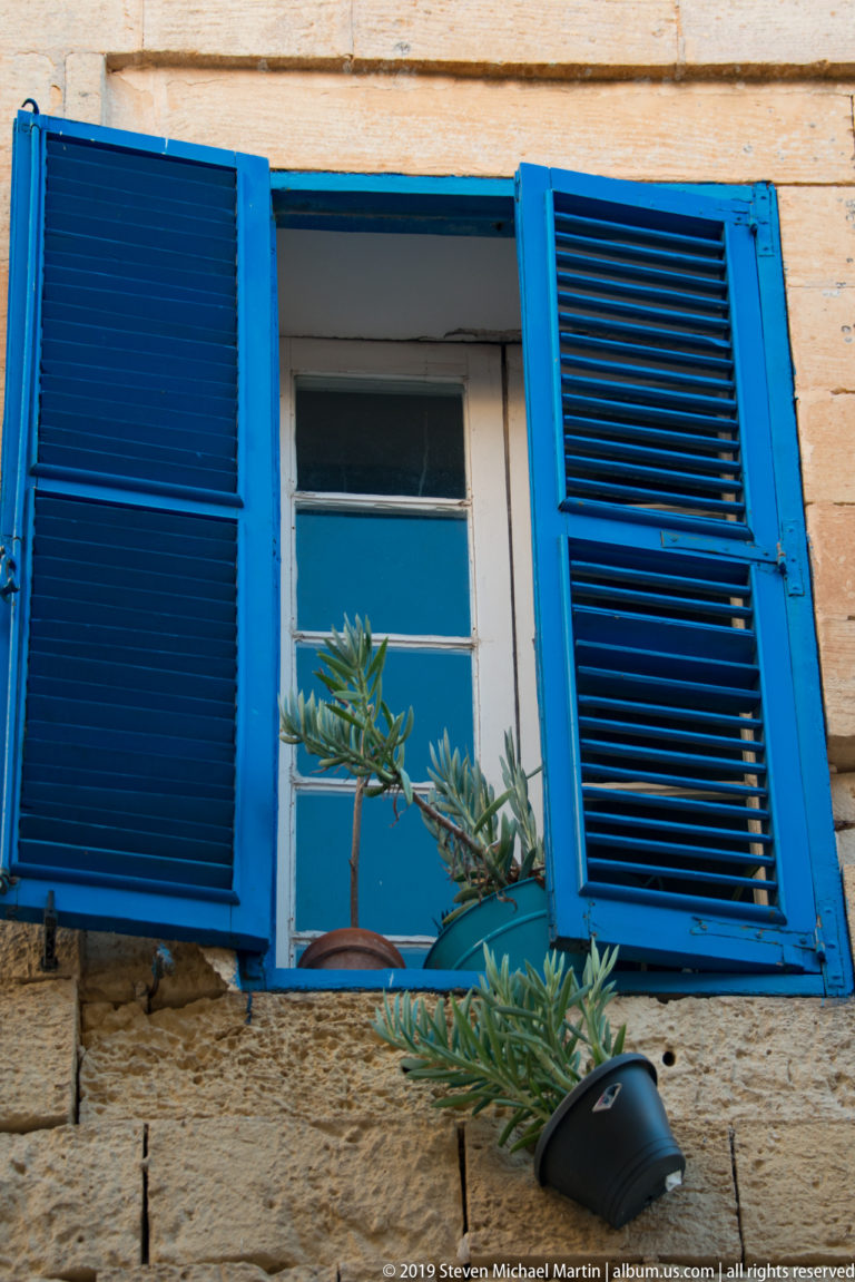 Bright blue louvered shutters opened around a window in a limestone wall, with leafy branch below.