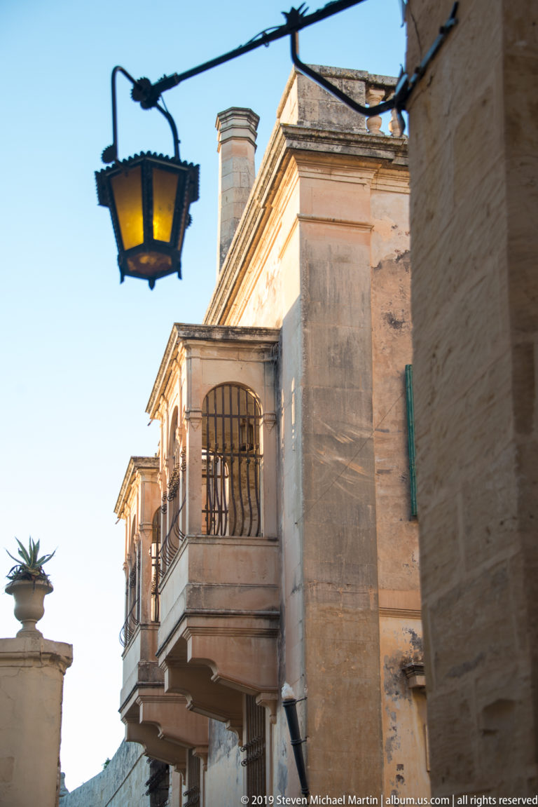 Limestone street corner with projecting balconies and a hanging lantern against a clear blue sky.