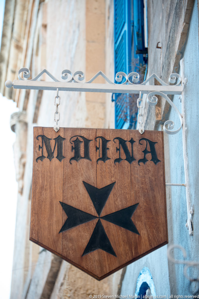 Wooden hanging sign reading MDIENA with a black Maltese cross on a limestone street façade.