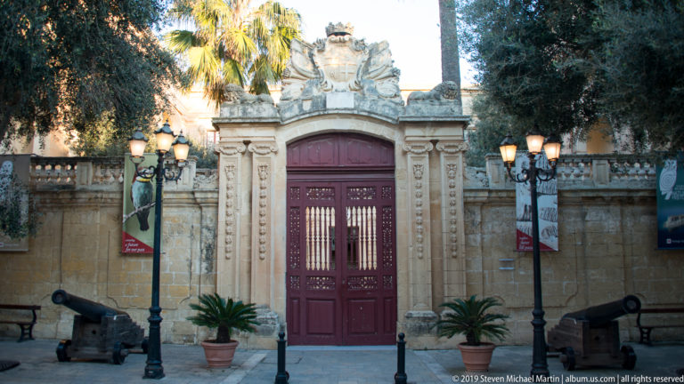 Ornate red double gate in a carved limestone portal, flanked by cannon, lamps, and potted palms.