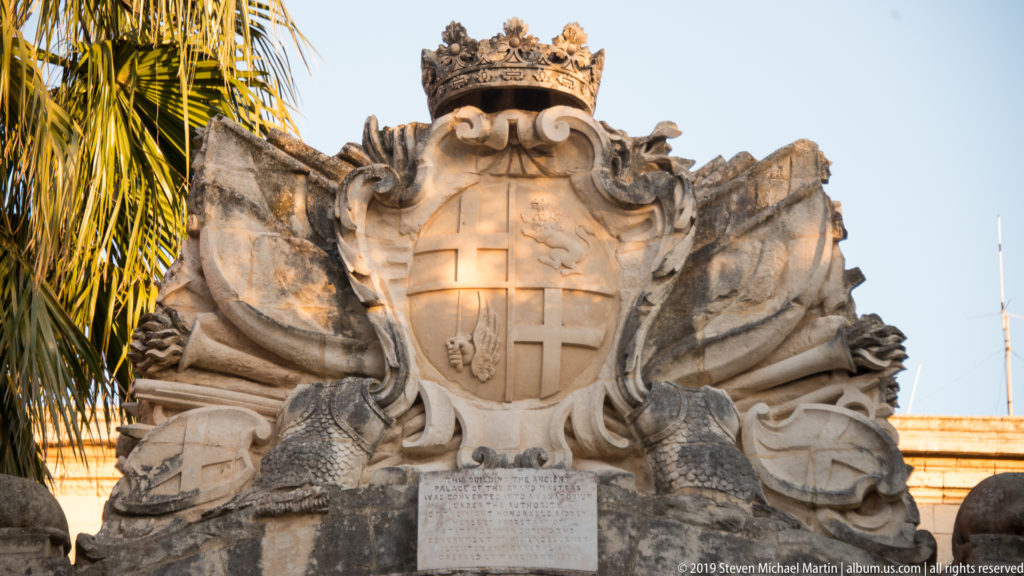 Large crowned stone coat of arms with a cross on the shield, surrounded by carved scrollwork.