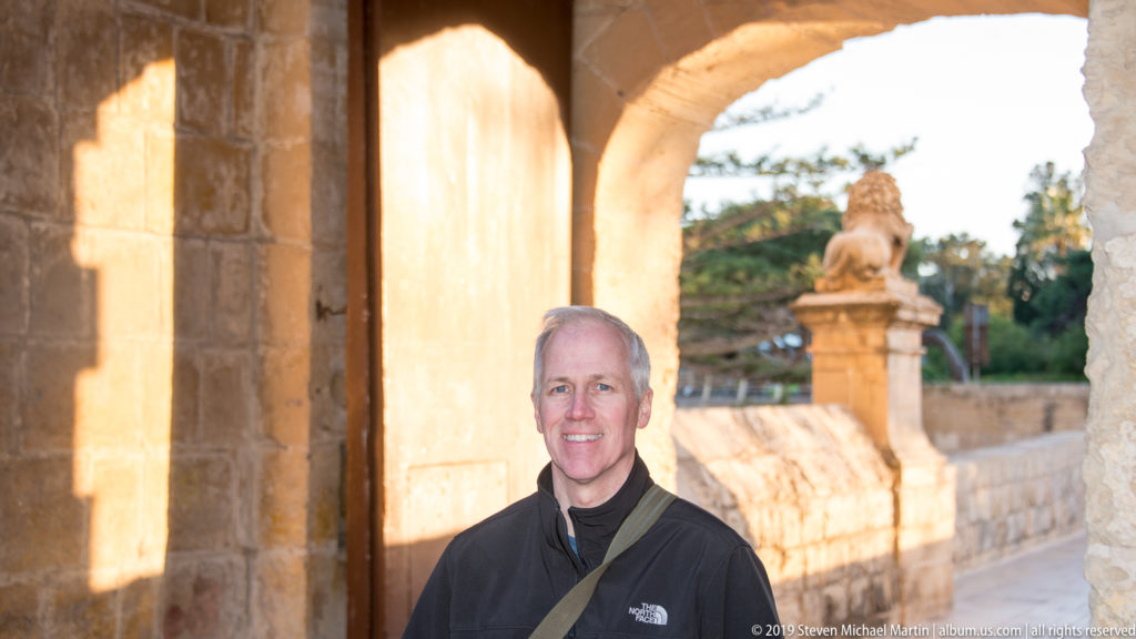 Smiling visitor in a dark jacket beneath a limestone arch, with a stone lion on a pedestal behind.