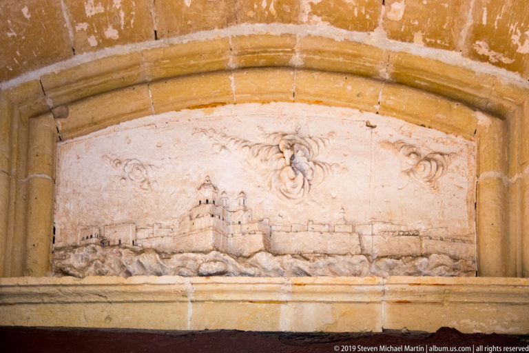 Weathered stone relief of a fortified skyline beneath decorative motifs within a rounded niche.