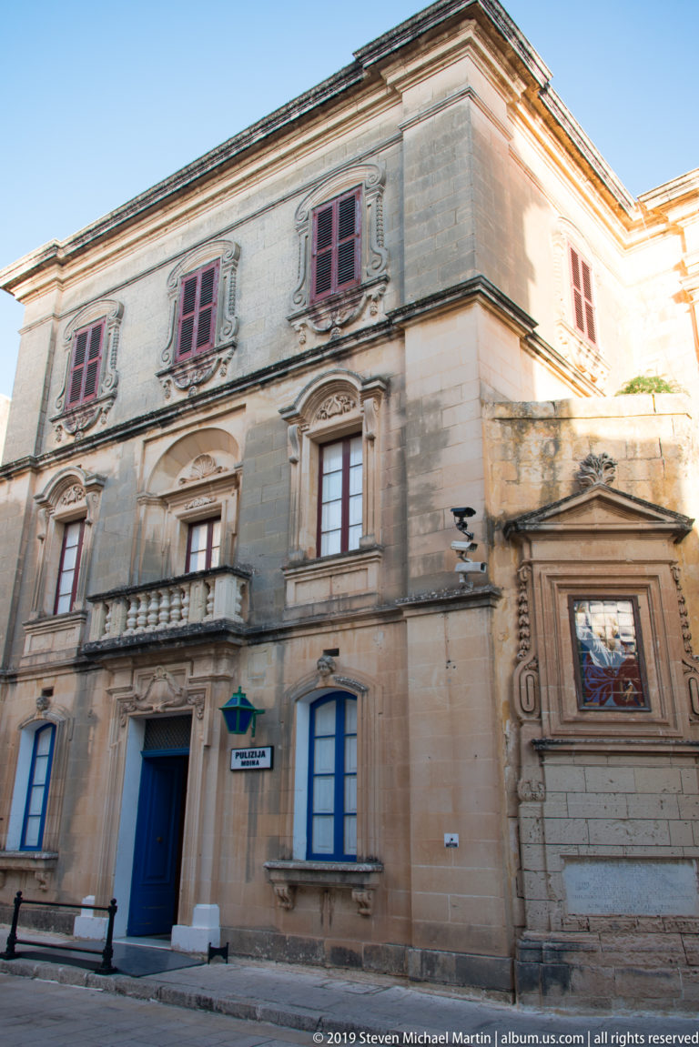 Corner limestone townhouse with carved balconies, red and blue shutters, tall windows, blue door, and a framed religious image on the side wall.