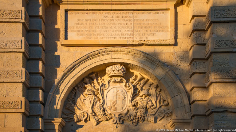 Carved coat of arms beneath a Latin inscription over the stone arch of Mdina Gate.