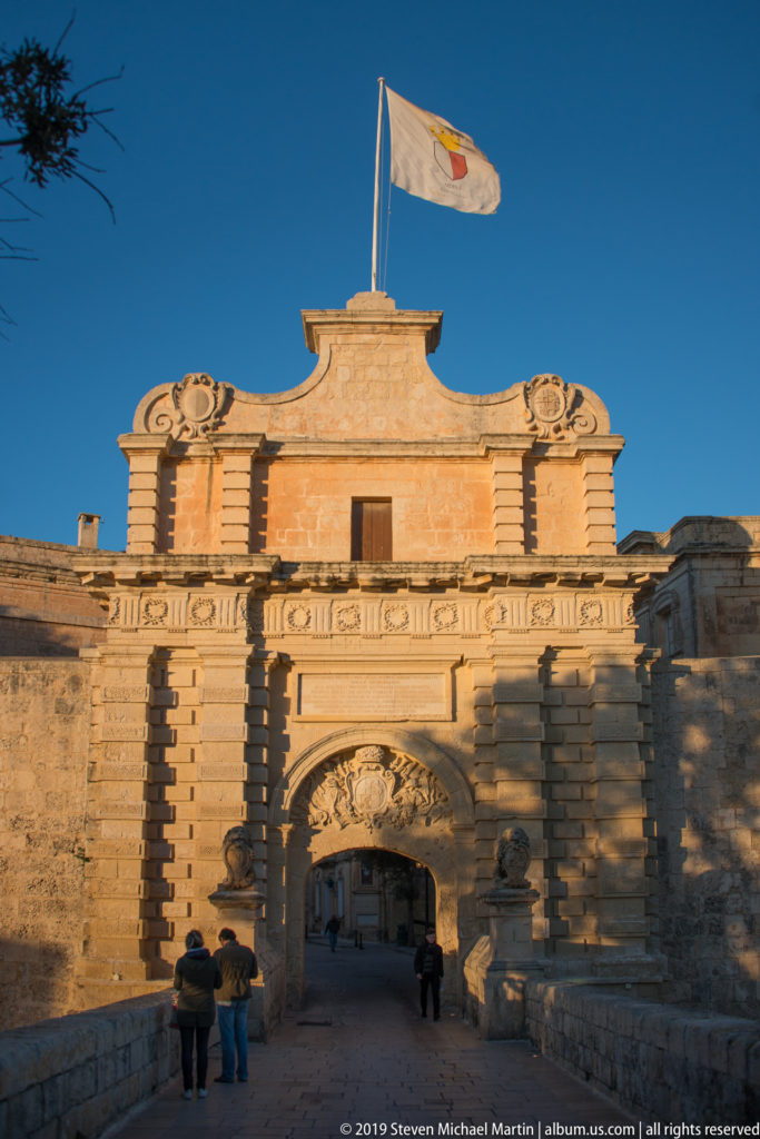 Baroque Mdina Gate with rusticated stonework, central arch, sculpted crest, Maltese flag, and visitors below.