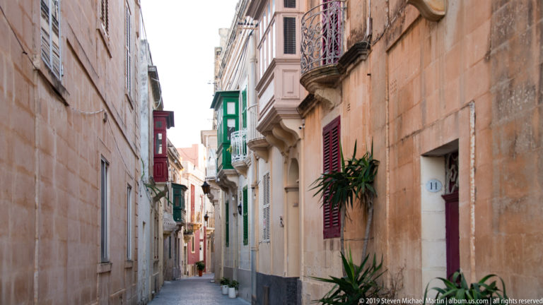 Narrow M’dina street lined with limestone façades, colorful shutters, projecting balconies, and potted plants.