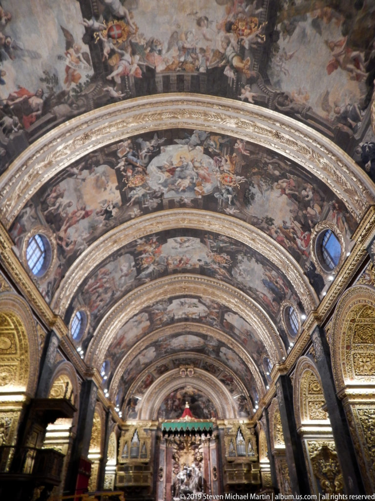 Upward view along a painted barrel vault divided by gilded ribs, with small windows and richly ornamented arches below.