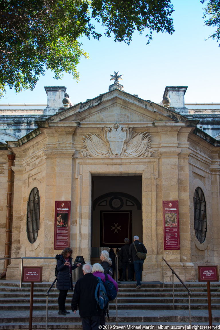 Visitors climb steps to St. John’s Co-Cathedral entrance with limestone pediment, heraldic carving, Maltese cross, and flanking oval windows.