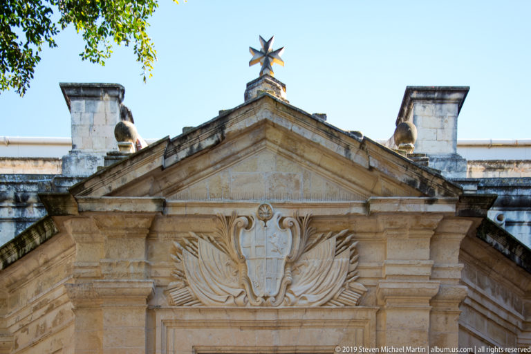 Limestone entrance pediment of St. John’s Co-Cathedral with central heraldic carving and a Maltese cross finial against blue sky.