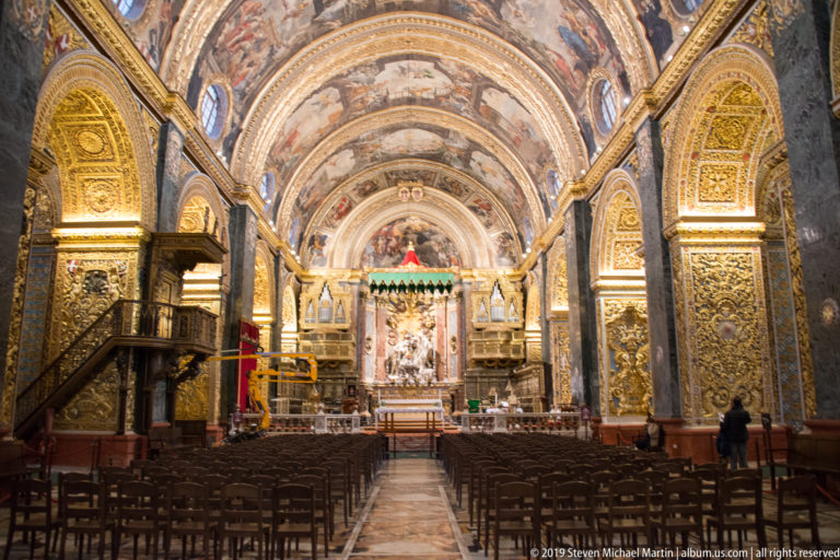 Central nave view toward the illuminated sanctuary beneath a painted vault, with pulpit at left and rows of chairs.