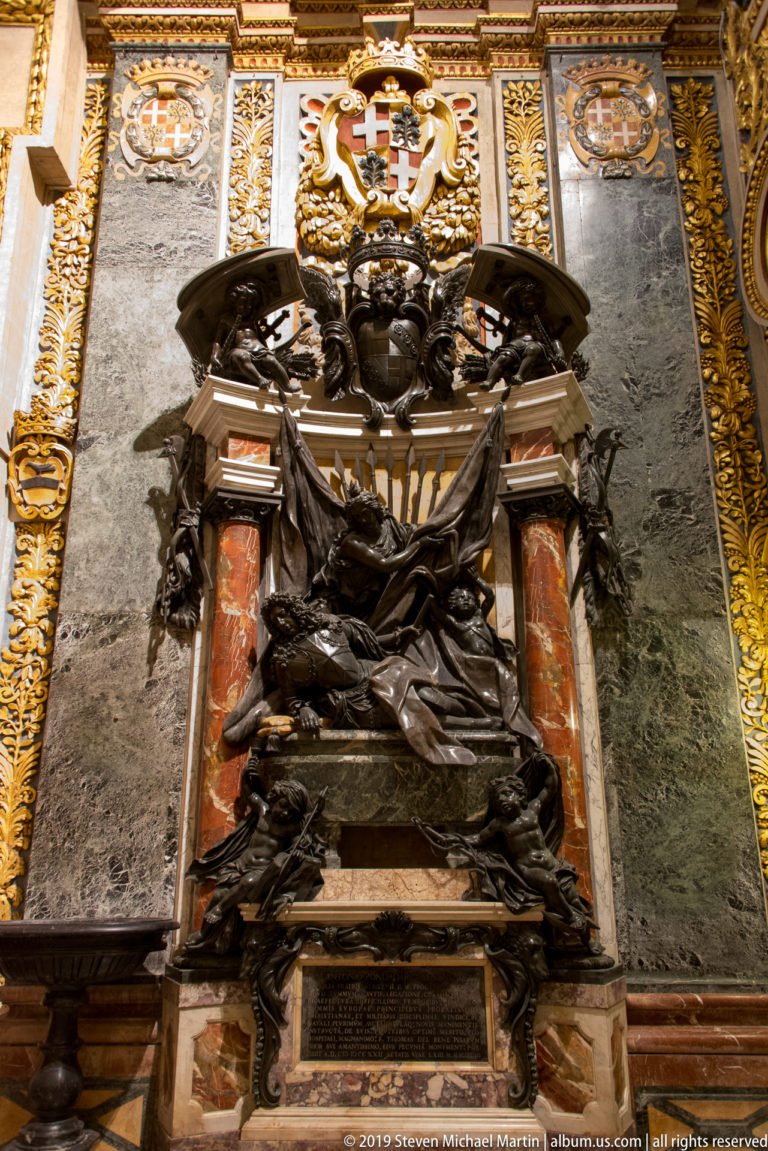 Dark draped sculptural figure in a memorial framed by reddish marble columns, with heraldic shields and gilded borders above.