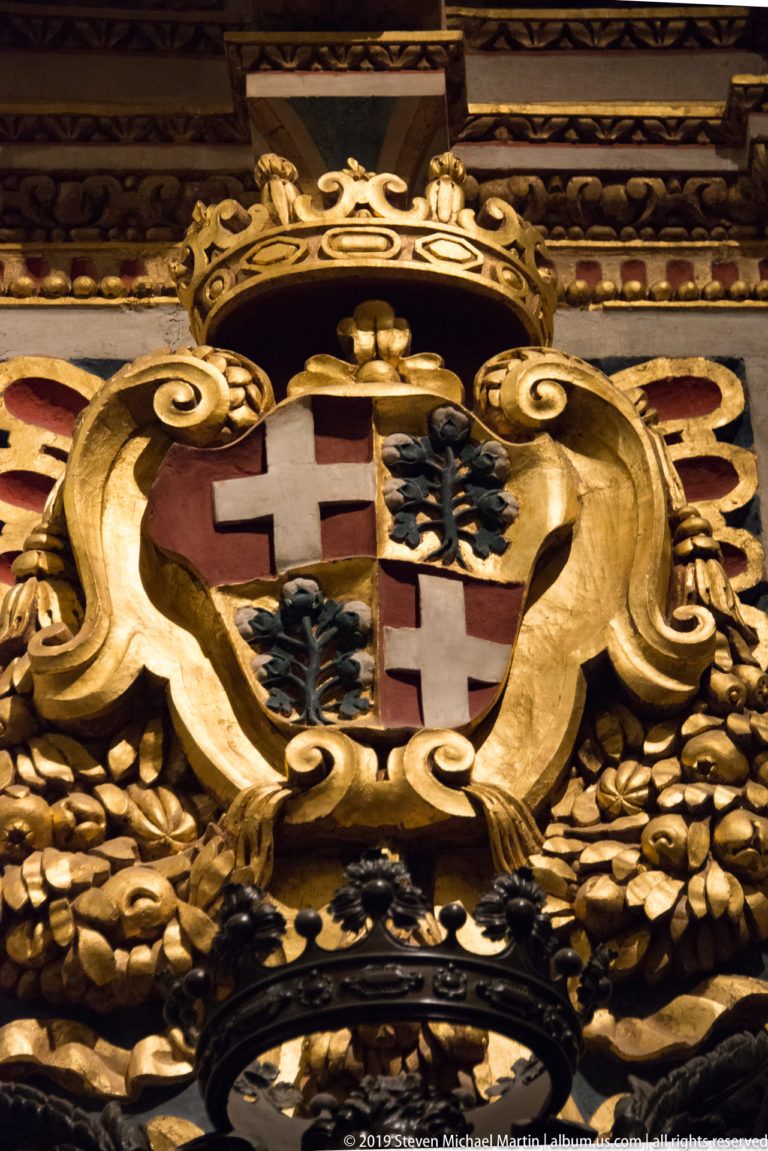 Gilded carved heraldic shield with red crosses and dark tree emblems surrounded by dense scrolling ornament.