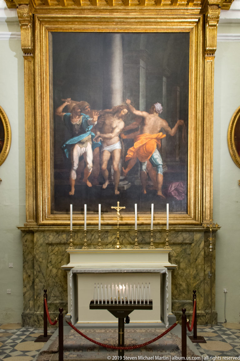 Large dark-toned religious painting in a gilded frame above a marble altar with candles and a low rail.