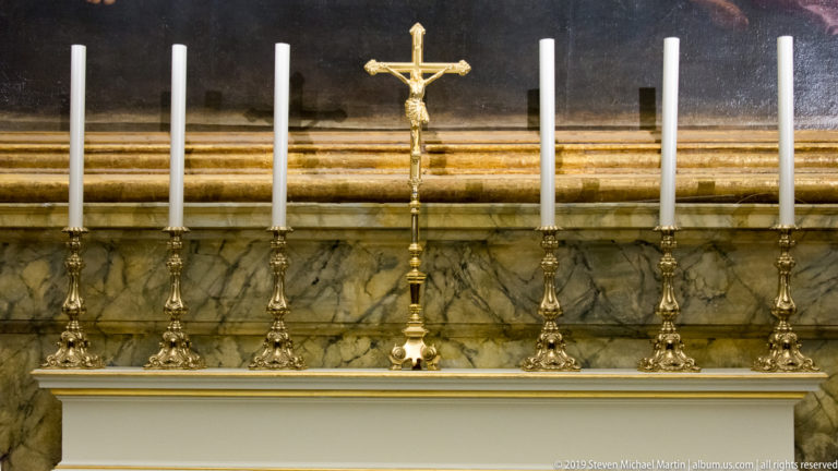Gold crucifix centered between six tall white altar candles in ornate holders against veined marble.
