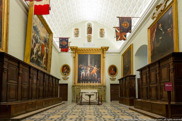 Axial view between dark wooden stalls toward a large end-wall painting, with flags and smaller portraits in a white chamber.