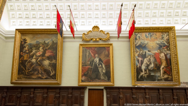 Three framed paintings above dark paneling, with a central portrait flanked by battle scenes and heraldic flags overhead.