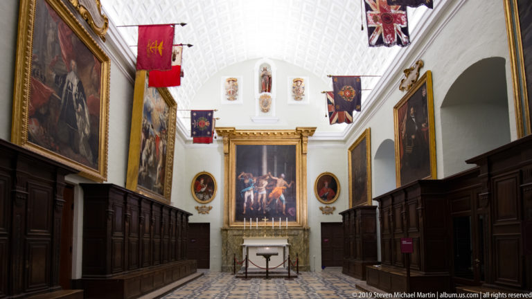 Bright white chamber with dark wooden stalls, large paintings, hanging heraldic flags, and a small altar-like table.