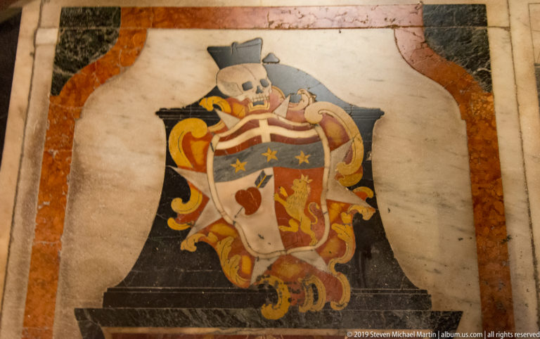 Colorful inlaid coat of arms with helmet, mantling, stars, and animal device set into pale marble flooring.